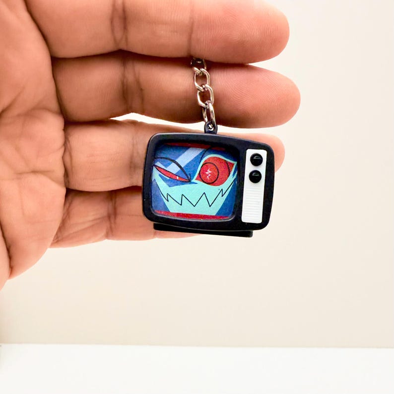May include: A black and white TV-shaped keychain with a cartoon face on the screen. The screen displays a blue background with a red eye and a jagged, toothy grin. The keychain is held by a silver chain.