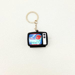 May include: A black keychain featuring a small, retro-style television. The TV screen displays a cartoon face with red eyes and a jagged white grin against a blue background. The keychain has a silver ring and chain.