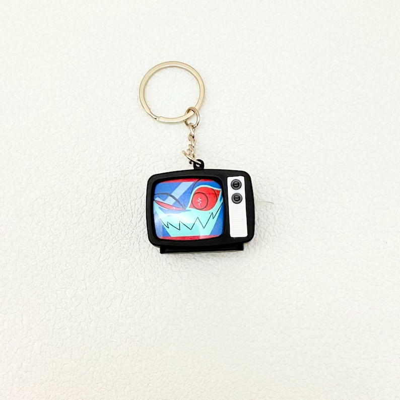 May include: A black keychain featuring a small, retro-style television. The TV screen displays a cartoon face with red eyes and a jagged white grin against a blue background. The keychain has a silver ring and chain.