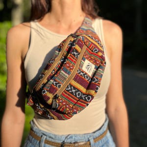 May include: A bum bag featuring a multi-coloured woven pattern in shades of red, yellow, black, and blue. The bag has two zipped compartments with tan trim and a small rectangular white label with the text Everest on the front panel.