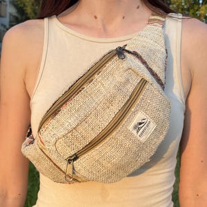 Pode incluir: Uma pochete tecida em bege com acabamento em tecido texturizado, apresentando dois compartimentos com zíper e um pequeno emblema retangular branco com o texto preto EVEREST.GEAR e um gráfico de montanha. A alça tem um padrão listrado colorido.