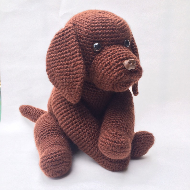 May include: A brown crocheted stuffed dog toy sitting on a white surface. The dog has black eyes and a brown nose.