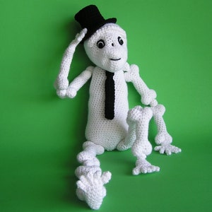 May include: A white crocheted skeleton wearing a black top hat and a black tie. The skeleton is sitting with its legs crossed and its arms resting on its knees.