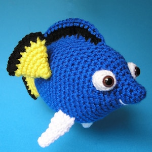 Può includere: Un pesce all'uncinetto blu con pinne gialle e nere e una coda bianca all'uncinetto. Il pesce ha grandi occhi bianchi con pupille marroni.