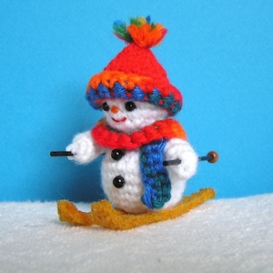 May include: A crocheted snowman wearing a red and orange hat with a blue and green scarf, holding ski poles and standing on skis.