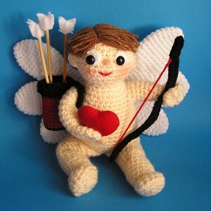 Puede incluir: Un amigurumi de crochet que representa a un cupido con cabello castaño, sosteniendo un corazón rojo y un arco y flecha. El cupido lleva un halo blanco de crochet y tiene alas.