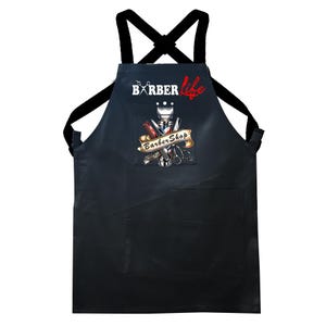 Può includere: Grembiule da barbiere nero con tasca e spalline nere. Il grembiule presenta la scritta "BARBER life" in bianco e rosso, insieme a una grafica di strumenti da barbiere e le parole "Barber Shop".