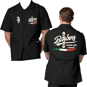 Peut inclure: Chemise de barbier noire à fermeture éclair et manches courtes. Le mot "Barbers" est écrit en script, avec un motif de poteau de barbier et les mots "Barber Shop Haircut". Le dos de la chemise affiche également le logo du salon de coiffure.