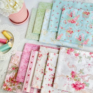 May include: A collection of floral fabric squares in various pastel colors, including pink, green, and blue. Some fabrics feature rose patterns, while others have polka dots or stripes. A pink flower pot with a white flower and spools of thread are also visible.