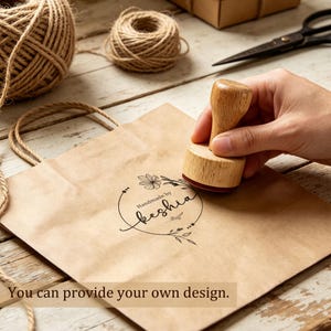 Könnte beinhalten: Ein runder Holzstempel mit Griff, der ein schwarzes Tintenmotiv auf eine braune Papiertüte drückt. Das gestempelte Design zeigt einen kreisförmigen Blumenkranz mit dem Text "Handmade by Keshia" sowie dem Text "You can provide your own design."