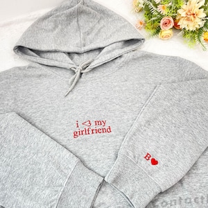 May include: A gray hoodie with a drawstring hood and long sleeves. The text "i <3 my girlfriend" is embroidered in red on the front. A red heart and the letter "B" are embroidered on the left sleeve.