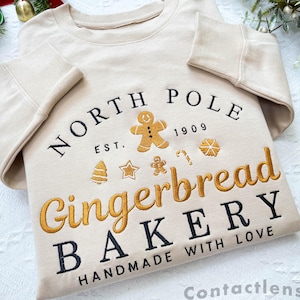 Może przedstawiać: Beżowa bluza z haftem "North Pole Gingerbread Bakery Est. 1909 Handmade with Love" w kolorze czarnym i złotym. Wzór zawiera piernikowego ludzika, laskę cukrową, gwiazdę, choinkę i płatek śniegu.