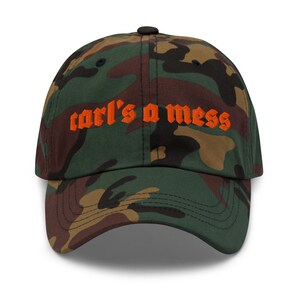 Puede incluir: Gorra de béisbol de camuflaje con el texto "carl's a mess" bordado en naranja. La gorra tiene una visera curva y un botón negro en la parte superior. El patrón del sombrero incluye tonos de verde, marrón y beige.