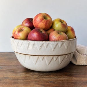 May include: A large, off-white ceramic bowl with a textured, repeating triangular pattern around the exterior, filled with a variety of red and yellow apples. The bowl features a horizontal brown line detail and a matte, slightly speckled finish.