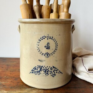 May include: A beige stoneware crock with two side handles, featuring a blue stamped number 4 inside a floral wreath and a blue floral design below. The crock is filled with a collection of various wooden rolling pins of different shapes.