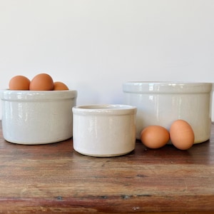May include: Three off-white ceramic crocks of varying sizes are arranged on a wooden surface. The largest crock contains several brown eggs. Two additional eggs sit beside the smallest crock. The crocks have a rustic, farmhouse aesthetic.