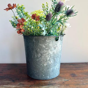May include: A galvanized metal bucket filled with a variety of colorful faux flowers. The arrangement includes rust-colored cosmos, purple thistles, white daisies, and various green foliage. The bucket sits on a wooden surface.