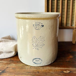 May include: A vintage, off-white stoneware crock with a tan rim. It features a blue number "5" and a maple leaf design. A wooden washboard and a folded cloth are in the background. The crock has a "MITCHELL STONEWARE COMPANY" stamp.