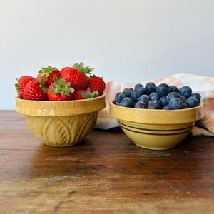 May include: Two yellow ceramic bowls filled with fresh fruit. One bowl holds ripe red strawberries, and the other is filled with blueberries. The bowls sit on a wooden surface, with a white background.