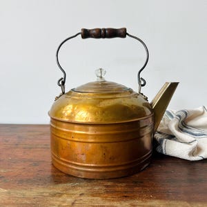 May include: A vintage copper tea kettle with a polished brass-toned lid and body. The kettle features a dark brown wooden handle, a clear glass knob on the lid, and a sturdy metal spout. The surface shows natural patina and subtle metallic textures.