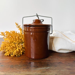 May include: A cylindrical brown glazed ceramic crock with a matching lid and a gray metal wire bail handle. The container features a smooth, glossy finish and a simple, functional design, standing upright with a secure lid and a sturdy metal handle.