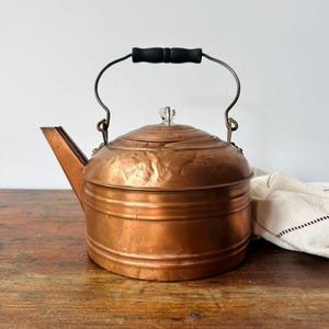 May include: A copper tea kettle with a cylindrical body, a straight spout, and a hinged metal handle featuring a black wooden grip. The lid is topped with a small, clear glass knob. The metal surface shows signs of wear and natural patina.