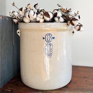 May include: A large, off-white stoneware crock with blue lettering and a graphic. The crock is filled with cotton stems. The crock has two small handles on the sides. The crock is sitting on a wooden surface.