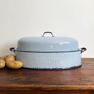 May include: A light blue enamelware roasting pan with a lid and black trim. The word "SAVORY" is embossed on the side. The pan sits on a wooden surface with potatoes nearby.
