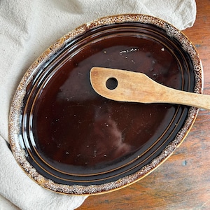 May include: Oval brown ceramic platter with a wooden spatula. The platter has a glossy finish and a textured, speckled border. The spatula is light brown with a hole in the handle. The platter is on a wooden surface.
