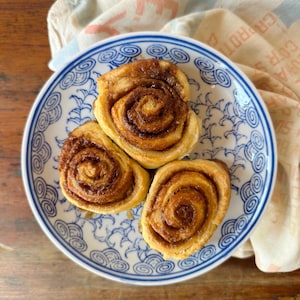 May include: Three golden-brown cinnamon rolls with visible swirls of cinnamon sugar filling arranged on a white ceramic plate featuring a blue and white stylized wave pattern. The pastry dough is baked to a light brown color with dark brown sugar.