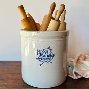 May include: A cylindrical off-white stoneware crock featuring a blue maple leaf logo with the text 3 WESTERN STONEWARE CO. MONMOUTH ILL. The crock is filled with several wooden rolling pins of varying shades and handle designs.