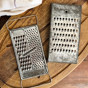 May include: Two vintage metal graters on a wooden cutting board. One grater is rectangular with the text "ALL IN ONE" and the other is labeled "WONDER SHREDDER". The graters are silver and show signs of wear.