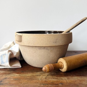 May include: A tan stoneware mixing bowl with a dark brown interior, a wooden spoon, and a wooden rolling pin rest on a wooden surface. A white and blue patterned cloth is partially visible.