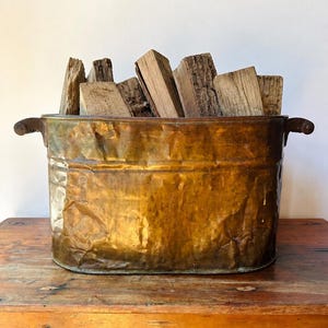 May include: An oval-shaped, hammered copper bucket with two side handles, filled with several pieces of rough-cut, light brown firewood. The metal container features a weathered, textured surface with visible dents and a warm, metallic finish.