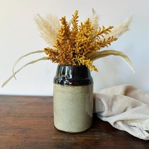 May include: A ceramic vase with a dark brown glazed neck and a light beige crackled body, containing a dried floral arrangement of golden-yellow textured stems and fluffy cream-colored pampas grass plumes.