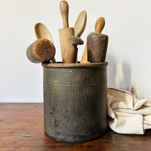 May include: A dark, weathered cylindrical stoneware crock filled with an assortment of vintage wooden kitchen utensils, including various spoons, a small rolling pin, and a wooden pestle, all showing signs of age and use.