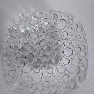 Peut inclure: Bol en verre transparent avec une surface texturée ornée de nombreuses formes en forme de bulles. Le bol est de forme carrée avec des coins arrondis et un bord légèrement évasé. Le verre transparent laisse passer la lumière.