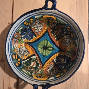 May include: A white ceramic bowl with blue handles and a colourful floral design. The bowl features a geometric pattern with blue, green, yellow, and orange colours.
