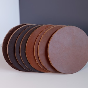 May include: A collection of round leather coasters in various shades of brown, from dark espresso to light tan. The coasters are stacked, showcasing their circular shape and natural texture. The edges are slightly raised.