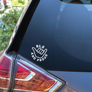 May include: White vinyl sticker with the text "RLS AND PROUD" in a circular design featuring a hand gesture. The sticker is on the rear window of a car, near the rear light.