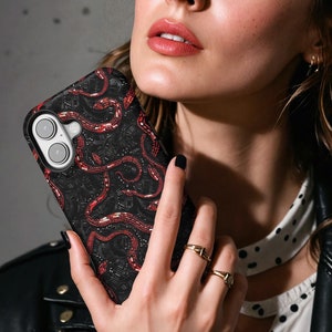 Könnte beinhalten: Schwarze Handyhülle mit rotem Schlangenmuster. Die Hülle hat eine strukturierte Oberfläche und einen Kameraausschnitt. Die Handyhülle wird vor einer Person gehalten, die eine schwarze Lederjacke und ein weißes Polka-Dot-Hemd trägt.