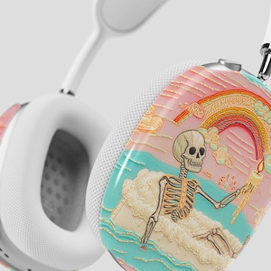 May include: White over-ear headphones featuring a decorative skin on the ear cups. The design depicts a skeleton sitting on a cloud, holding a lit candle, with a rainbow, pink sky, and turquoise water in the background.
