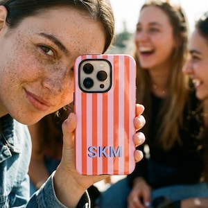May include: A rectangular smartphone case featuring a vertical striped pattern in alternating shades of coral orange and light pink. The center of the case displays the capital letters SKM in a dark blue, bold, sans-serif font with a drop shadow effect.