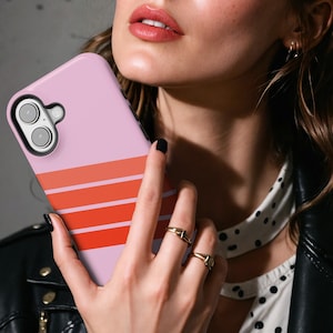 May include: A light pink smartphone case featuring three horizontal stripes in varying shades of orange and red. The case has a black camera cutout frame and covers the back of the device, showcasing a smooth finish and a clean, modern aesthetic.