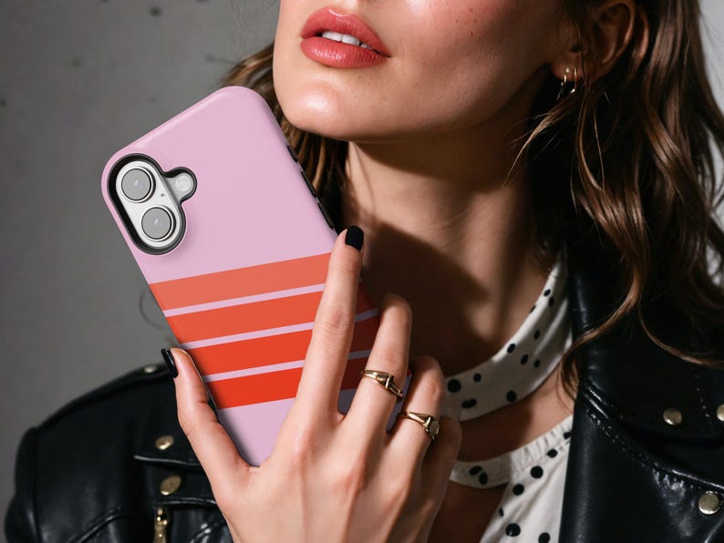 May include: A light pink smartphone case featuring three horizontal stripes in varying shades of orange and red. The case has a black camera cutout frame and covers the back of the device, showcasing a smooth finish and a clean, modern aesthetic.