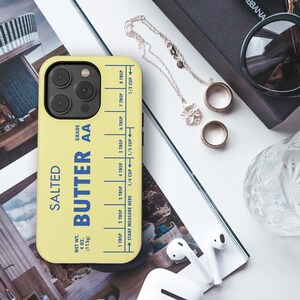 May include: A yellow phone case with a butter stick design. The case has a blue and white label that reads "Salted Butter AA" and includes a measurement guide for tablespoons and cups. The case is designed for an iPhone.