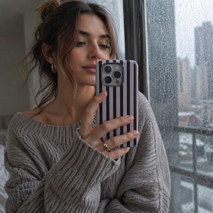 May include: A smartphone with a striped case in shades of white and burgundy is held up in front of a window. The case has vertical stripes. The person holding the phone is wearing a gray sweater and gold hoop earrings.