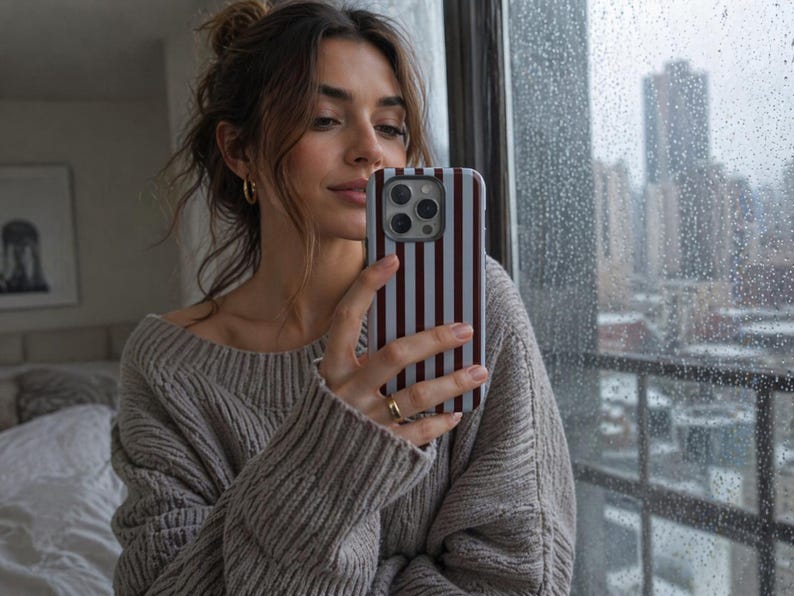 May include: A smartphone with a striped case in shades of white and burgundy is held up in front of a window. The case has vertical stripes. The person holding the phone is wearing a gray sweater and gold hoop earrings.
