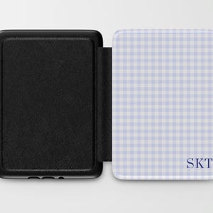May include: Black e-reader cover with a textured finish, open to reveal a light blue and white gingham pattern. The cover features the initials "SKT" in a dark blue font. The e-reader cover is designed to protect and personalize the device.