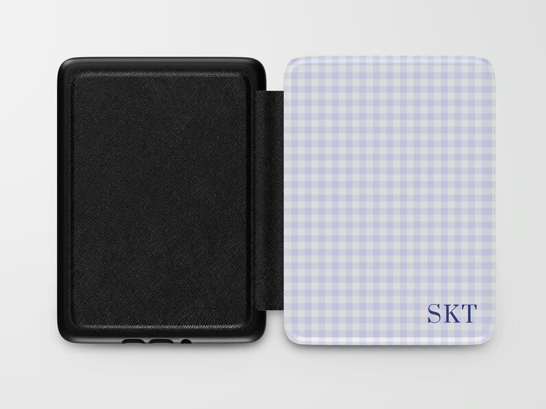 May include: Black e-reader cover with a textured finish, open to reveal a light blue and white gingham pattern. The cover features the initials "SKT" in a dark blue font. The e-reader cover is designed to protect and personalize the device.